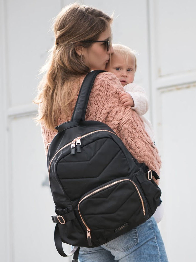storksak hero quilt black, changing bag backpack, in quilted nylon with rose gold hardware, mum wearing bag and holding baby