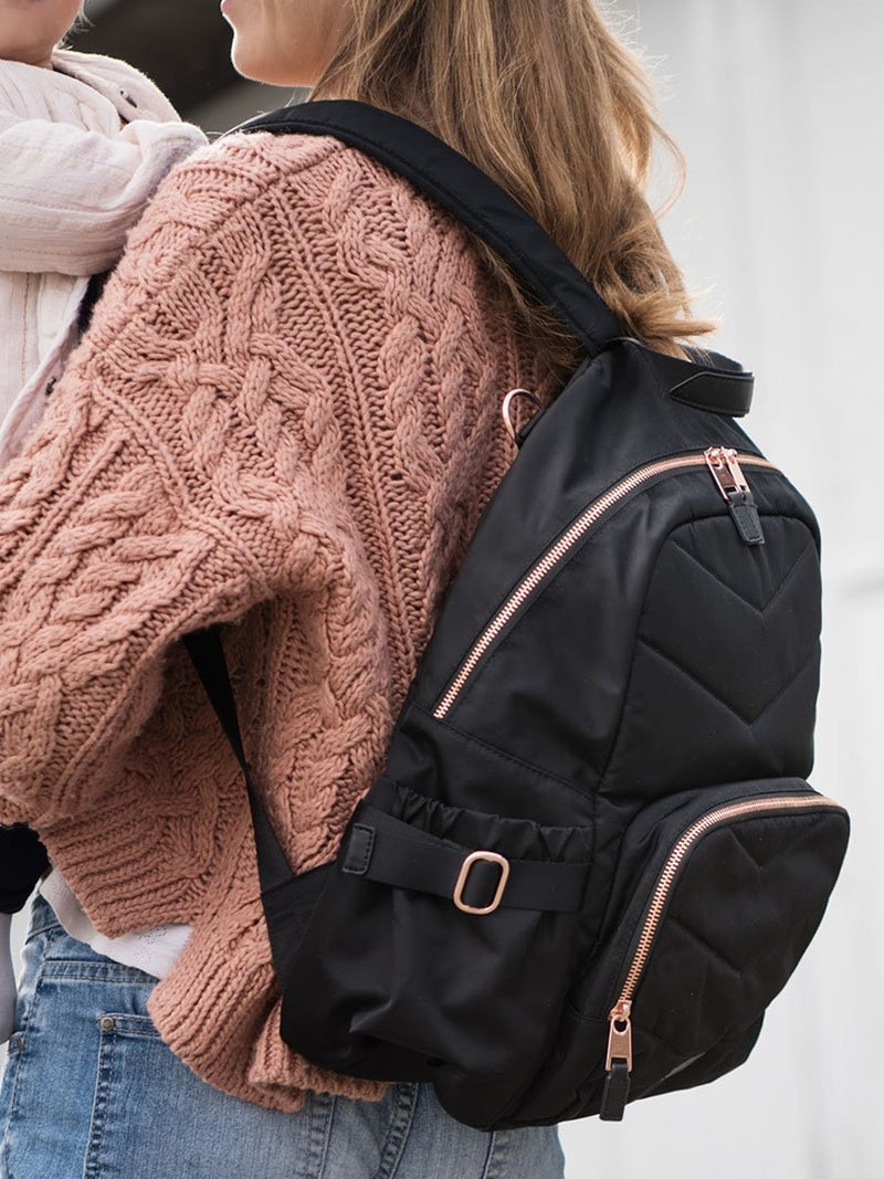 storksak hero quilt black, changing bag backpack, in quilted nylon with rose gold hardware, side view of mum wearing bag and holding baby